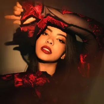 A woman with long dark hair poses with one arm raised, wearing a sheer black top with red floral patterns, lit by warm, dramatic lighting.