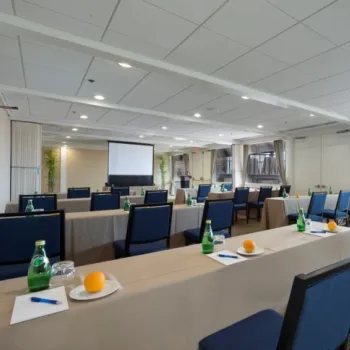 Conference room setup with rows of tables and blue chairs, each place setting has a water bottle, orange, pen, and paper; a projector screen is at the front.