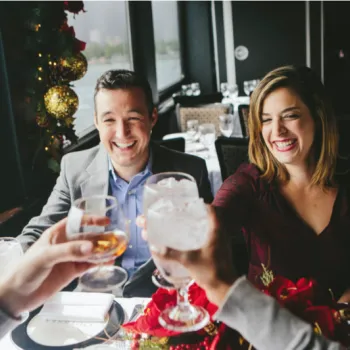 Two people sit at a restaurant table decorated with red flowers and raise glasses in a toast with others, smiling by a window with a festive holiday display.
