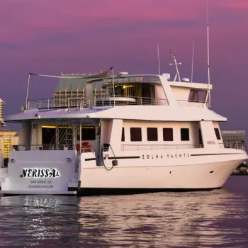 A white yacht labeled "Nerissa" from Zolna Yachts is docked on calm water at sunset with a city skyline in the background.