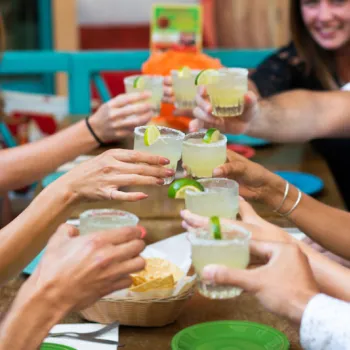 A group of people sit around a table outdoors, raising glasses of margaritas in a toast, with a basket of chips in the center.