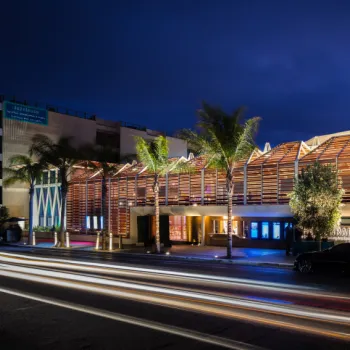 A modern building with wooden slats and illuminated exterior lighting, seen at night with palm trees and blurred car lights in the foreground.