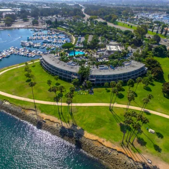 Aerial view of a curved waterfront hotel surrounded by green lawns, palm trees, walking paths, a marina with boats, and a pool area.