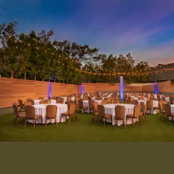Outdoor event space with round tables covered in white cloths and brown chairs, set up on artificial grass with string lights overhead at dusk.