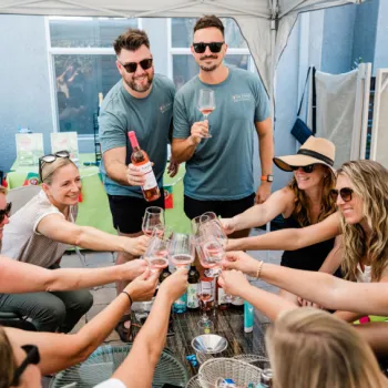 A group of people clink glasses of rosé wine around a table under a canopy, smiling and celebrating together.