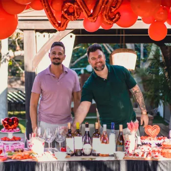 Two men stand behind a decorated table with wine and desserts, under red balloons and a "love" balloon, at an outdoor event.