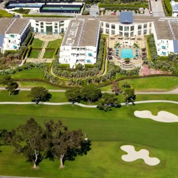 Aerial view of a large resort hotel with a central pool, surrounded by landscaped gardens, adjacent to a green golf course with sand bunkers and trees.