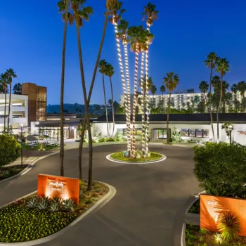 A hotel entrance at dusk with tall palm trees wrapped in lights, landscaped gardens, and a colorful mural on a nearby building.