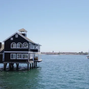 A black building on wooden stilts sits over calm blue water, with a white sailboat and a distant shoreline visible under a clear sky.