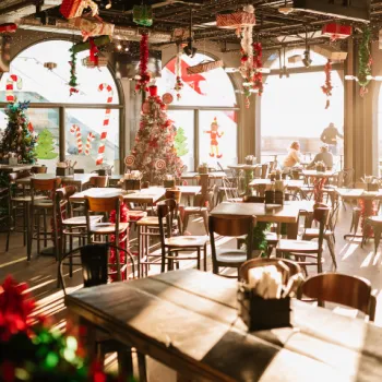 A festively decorated restaurant with Christmas trees, ornaments, and holiday-themed decorations, featuring empty tables and sunlight streaming through large windows.