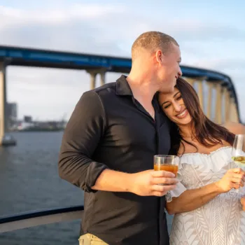 A couple stands on a boat holding drinks, with a bridge and city skyline in the background on a sunny day.