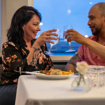 A woman and a man sit at a restaurant table, clinking wine glasses and smiling at each other, with plates of food and flowers on the table.