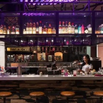 A bartender stands behind a modern bar with stools, mixing drinks; liquor bottles are displayed on a suspended shelf above the counter.