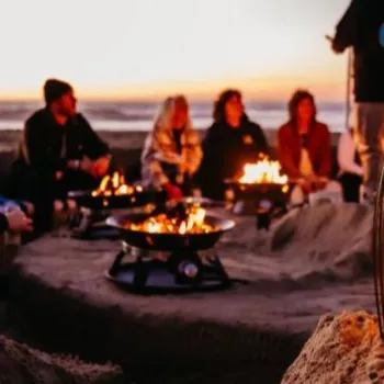 A group of people sit around multiple fire pits on a beach at sunset, with lanterns placed on the sand in the foreground.