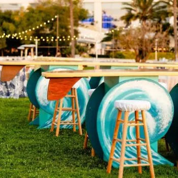 Outdoor event setup with high tables made from painted surfboards and barrels, surrounded by stools on a grass lawn with string lights and additional seating in the background.