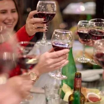 A group of people sit around a table, raising glasses of red wine in a toast, with food and bottles visible on the table.