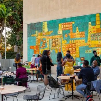 People gather in an outdoor courtyard with tables, eating and socializing near a large, colorful mural on the wall.