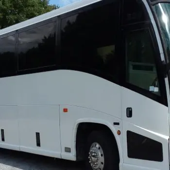 A large white coach bus is parked on a paved area near trees and grass under a partly cloudy sky.