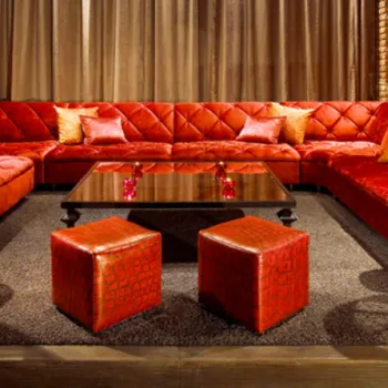 A red velvet sectional sofa with matching ottomans surrounds a dark coffee table in a room with exposed brick walls, modern lamps, and brown curtains.