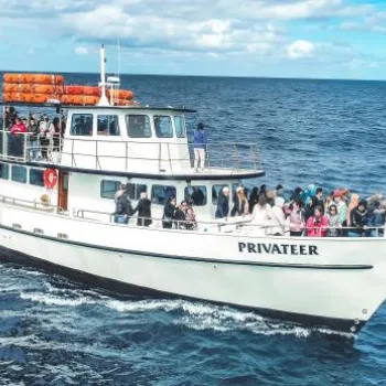 A white passenger boat named "Privateer" filled with people is traveling on the open ocean under a partly cloudy sky.