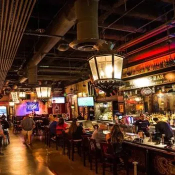 A dimly lit bar with patrons seated at the counter and tables, decorated with ornate lighting, vintage decor, and TV screens on the walls.