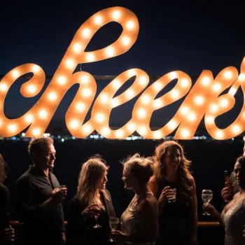 A group of people holding drinks stand in front of a large illuminated "cheers" sign at night.