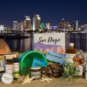 A variety of San Diego-themed gift items are arranged on sand at night, with the illuminated San Diego city skyline and waterfront in the background.