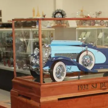 A model of a blue 1932 SJ Duesenberg car is displayed in a glass case in a museum with other exhibits and display cases in the background.