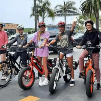 A group of people wearing helmets stand with their bicycles in a parking lot, with palm trees and buildings visible in the background.