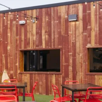 Outdoor patio with red chairs and black tables on artificial grass, string lights overhead, and a large chicken statue in the background near a wooden building.