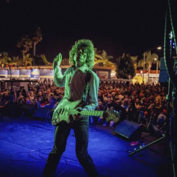 A guitarist performs on an outdoor stage at night, facing a large crowd, under colorful lighting with palm trees visible in the background.