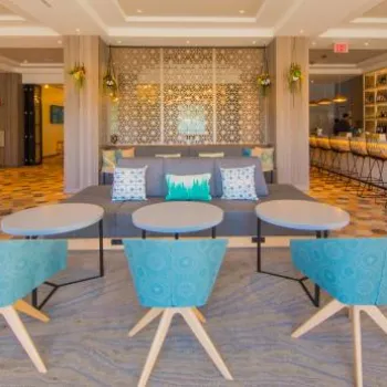 Modern hotel lobby with patterned tile flooring, blue chairs, round tables, a gray sofa, and a bar area with high stools in the background.