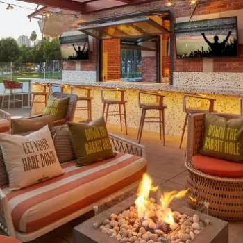 Outdoor lounge area with striped sofas, a fire pit, bar seating, and decorative pillows next to a bar with stools, set against an urban backdrop with palm trees and buildings.