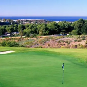 A golf course green with two sand bunkers overlooks a landscape of trees and shrubs, with buildings and the ocean visible in the distance under a clear sky.