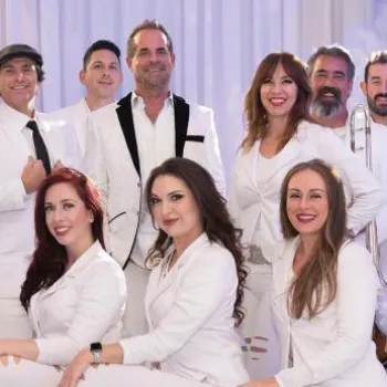 A group of musicians and singers dressed in white pose together, some holding instruments, against a light-colored backdrop.