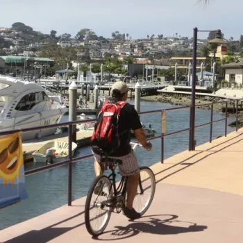 Two people ride bicycles on a waterfront pathway near docks and boats, with buildings and houses visible on a hillside in the background.