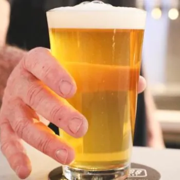 A person’s hand holding a pint glass filled with light beer, resting on a coaster on a bar counter.