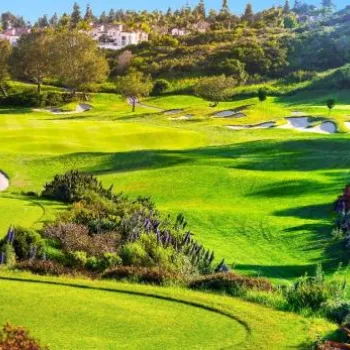 A scenic view of a well-maintained golf course with lush green fairways, sand bunkers, trees, and nearby residential buildings under a clear sky.