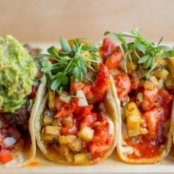 Five assorted tacos filled with various meats, vegetables, and toppings such as guacamole, salsa, herbs, and crumbled cheese, served on a wooden tray.