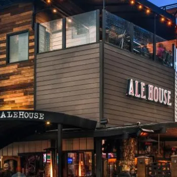 Pacific Beach Alehouse at dusk, featuring wood and metal exterior, large windows, outdoor string lights, and palm trees in the background.