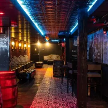 Dimly lit bar with red and blue accent lighting, barrel tables, patterned tile floor, stools along the bar, and vintage-style wall art and decor.