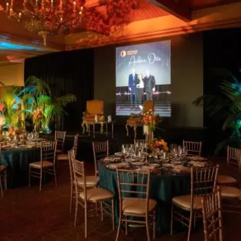 Ballroom set for a formal event with round tables, elegant place settings, floral centerpieces, and a stage with a screen and decorative plants.