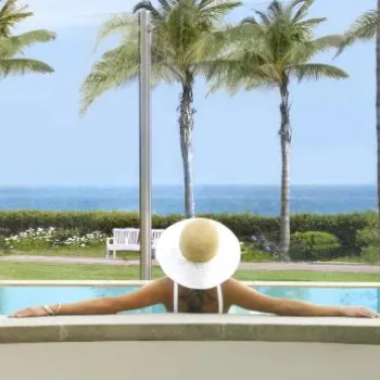 Person in a sunhat relaxes at the edge of an outdoor pool, facing the ocean, with palm trees and a lawn in the background and a pink towel on the poolside.