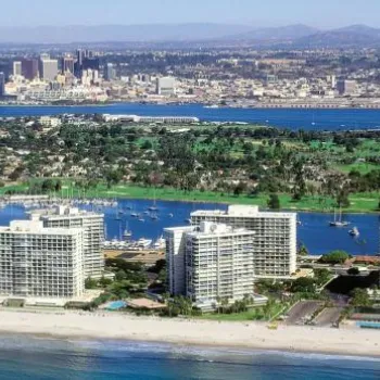 Aerial view of beachfront high-rise buildings, a marina, a green island, and a distant city skyline under a clear sky.