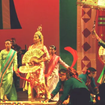 A group of performers in colorful traditional costumes act out a scene on stage with fans, umbrellas, and props under dramatic red lighting.
