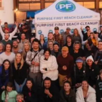 A large group of people pose together in front of a "Purpose First Beach Cleanup" tent on a sandy beach, with buildings visible in the background.