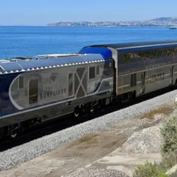 A modern Pacific Surfliner passenger train travels along tracks parallel to the coastline, with the ocean on one side and vegetation on the other.