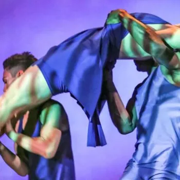 Two dancers in blue costumes perform a lift on stage under colorful lighting, with one dancer holding the other horizontally in the air.
