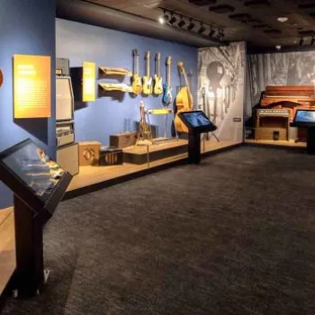 A museum exhibit displays various stringed instruments, including guitars and banjos, mounted on blue walls with informational panels and display cases.