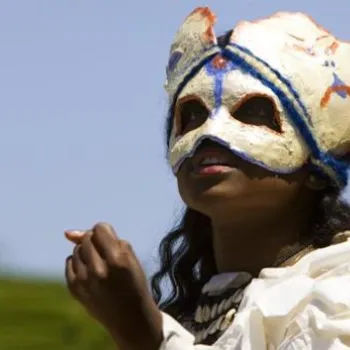 A person wearing a painted white mask and a white costume gestures with their hand against a clear sky background.
